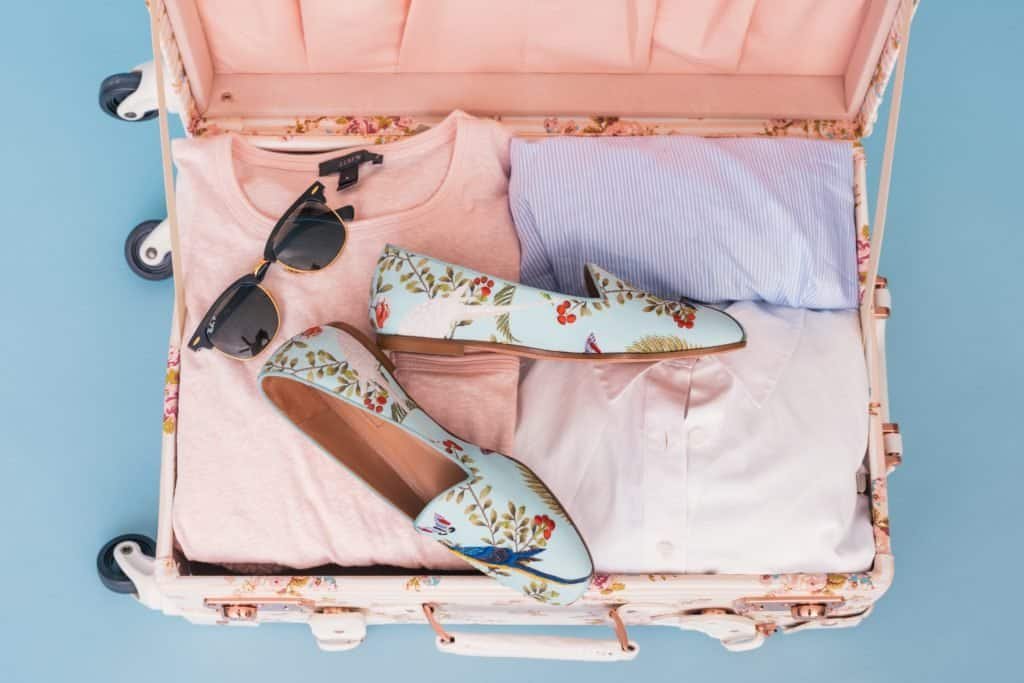 Pink Suitcase with clothes, sunglasses & shoes. Emphasizing where to buy travel essentials.